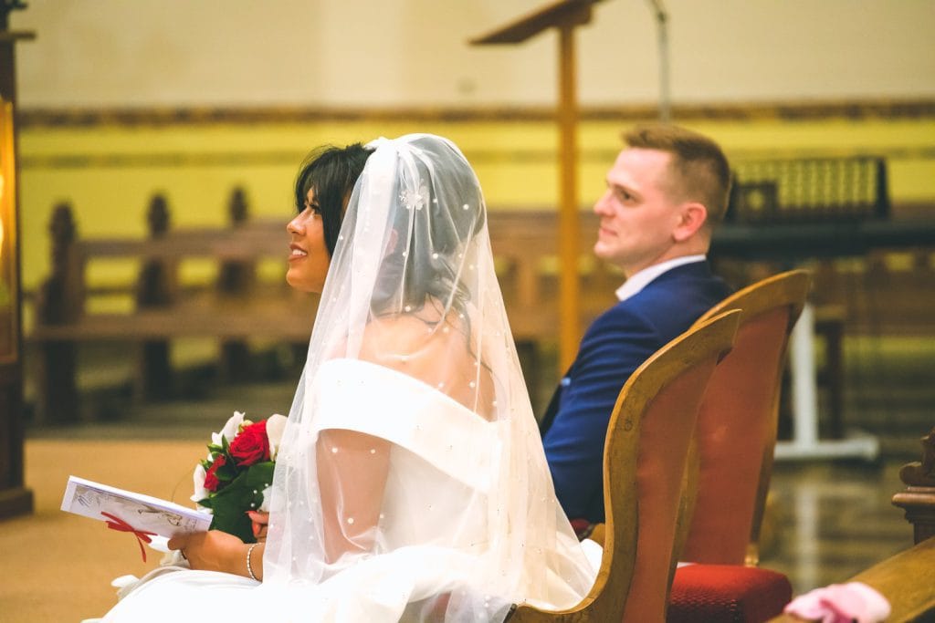 Portrait de profil d'un couple de mariés assis dans une église pendant leur cérémonie. La mariée porte un voile blanc orné de perles et tient un bouquet de roses rouges. Le marié, en costume bleu, regarde vers l'autel. Photo de mariage à Metz par Studio Salvucci