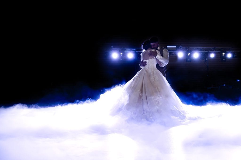 Couple de mariés enlacés pour leur première danse au milieu d'une fumée blanche épaisse au sol, créant un effet de nuages. Arrière-plan sombre avec des projecteurs de scène lumineux. Photographie de mariage par Studio Salvucci à Metz.