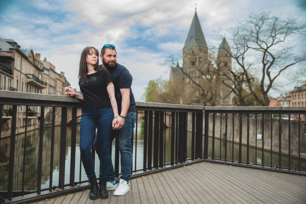 Un couple en tenue décontractée pose en se tenant la main sur une passerelle en bois à Metz. En arrière-plan, le Temple Neuf et les bâtiments historiques bordant la Moselle sous un ciel nuageux. Photographie lifestyle par Studio Salvucci