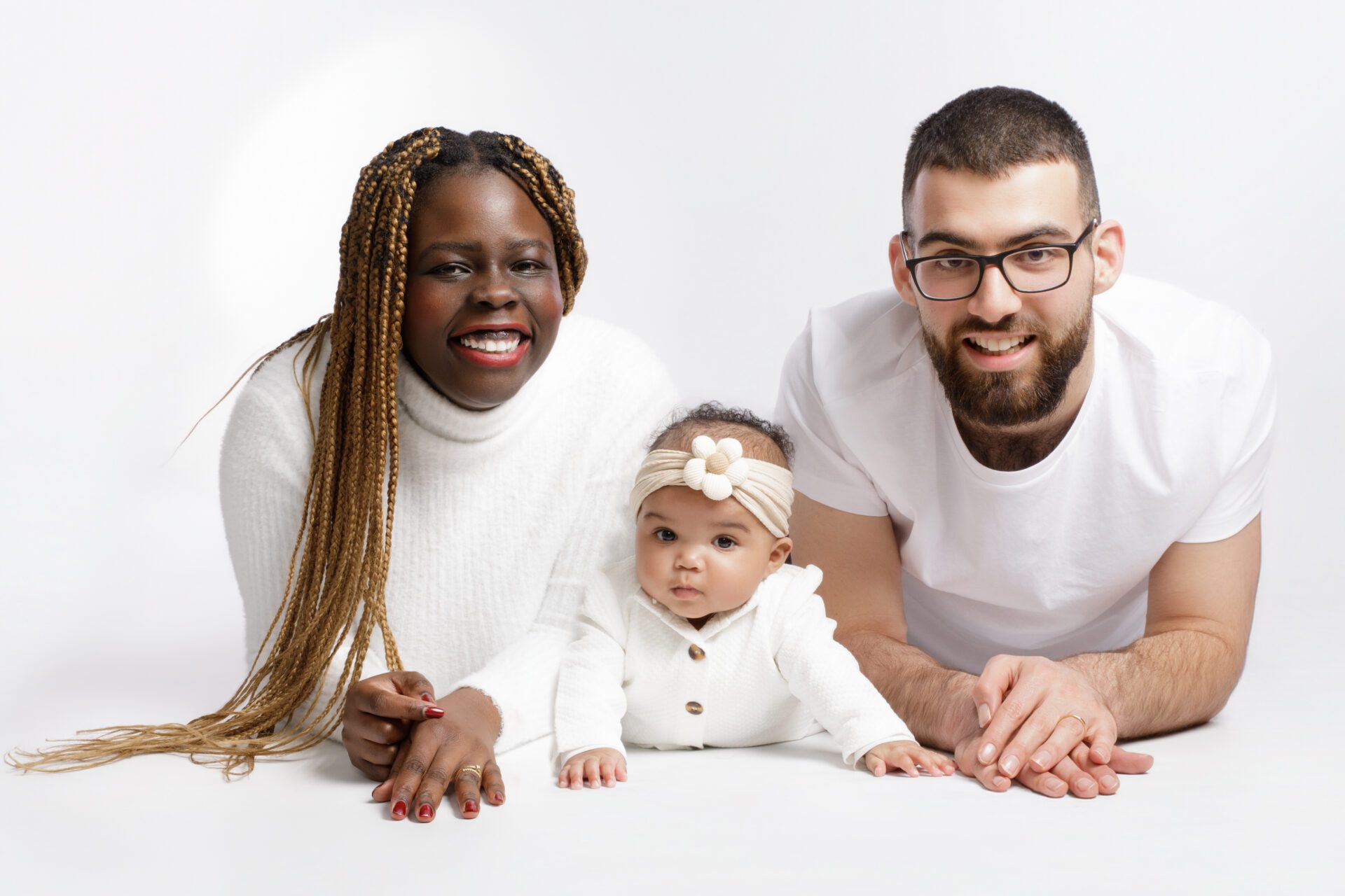 Portrait studio allongé d'une famille interraciale : mère noire souriante avec des tresses, père blanc barbu portant des lunettes, et leur bébé au centre sur le ventre avec un bandeau fleuri, tous vêtus de blanc sur fond blanc.