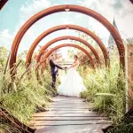 Couple de mariés se tenant par les mains au bout d'un chemin de bois sous une succession d'arches circulaires en métal rouillé, entouré de hautes herbes et fleurs. En arrière-plan, le clocher d'une église sous un ciel bleu. Photo par Studio Salvucci Metz