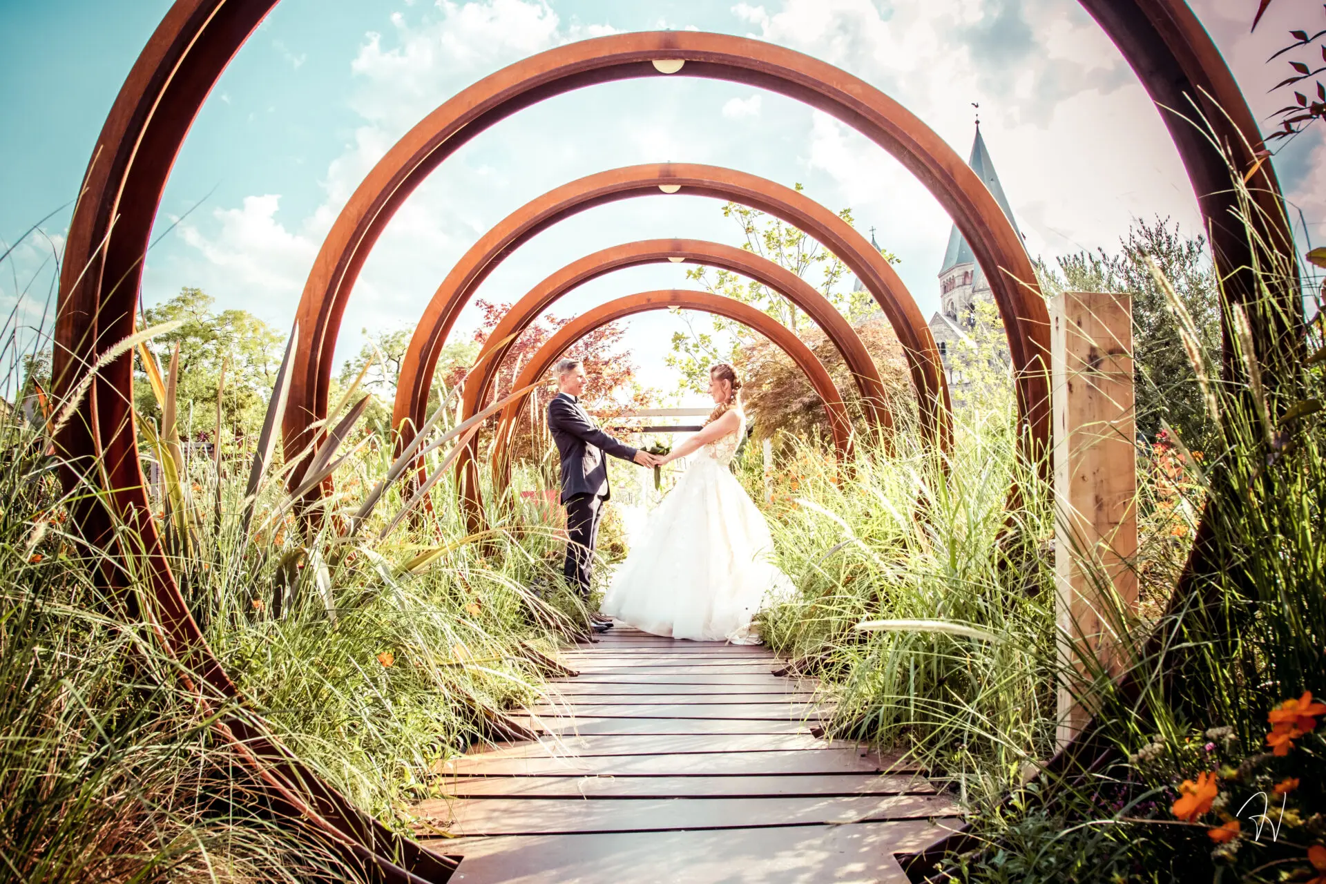 Couple de mariés se tenant par les mains au bout d'un chemin de bois sous une succession d'arches circulaires en métal rouillé, entouré de hautes herbes et fleurs. En arrière-plan, le clocher d'une église sous un ciel bleu. Photo par Studio Salvucci Metz
