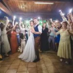 Un couple de mariés s'embrasse lors de leur première danse au centre d'une piste de danse. Ils sont entourés d'invités enthousiastes qui lèvent leurs téléphones portables pour éclairer la scène, créant un effet de lumières féeriques. Le lieu a un plafond en bois et le sol est en carrelage. Photographie de Studio Salvucci, Metz.