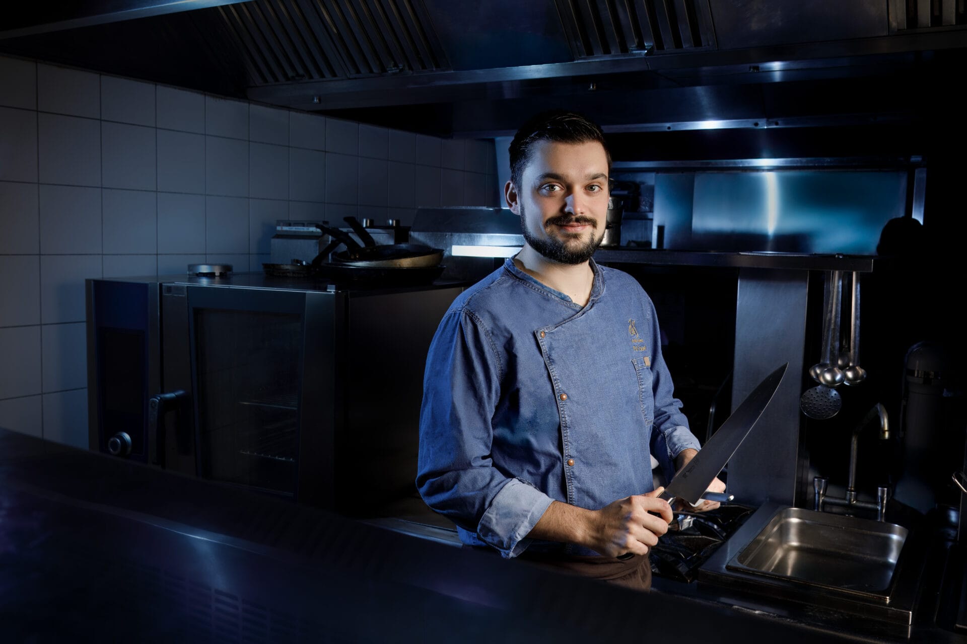 Jeune chef barbu dans une cuisine, tenant un grand couteau.