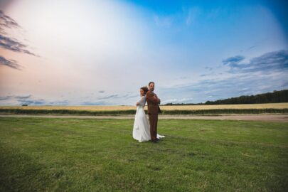 Dos à dos vers l'avenir : une parenthèse enchantée dans les champs de Moselle. Studio Salvucci, photographe de vos plus belles émotions à Metz et environs. Un couple de mariés debout dos à dos au milieu d'un vaste champ vert sous un ciel de fin de journée, l'homme portant un costume marron et la femme une robe de mariée blanche