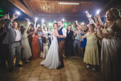 Une haie d'honneur lumineuse : la première danse comme un concert de lumières. Une soirée inoubliable capturée par Studio Salvucci, votre photographe d'émotions à Metz. Un couple de mariés s'embrasse lors de leur première danse au centre d'une piste de danse. Ils sont entourés d'invités enthousiastes qui lèvent leurs téléphones portables pour éclairer la scène, créant un effet de lumières féeriques. Le lieu a un plafond en bois et le sol est en carrelage. Photographie de Studio Salvucci, Metz.